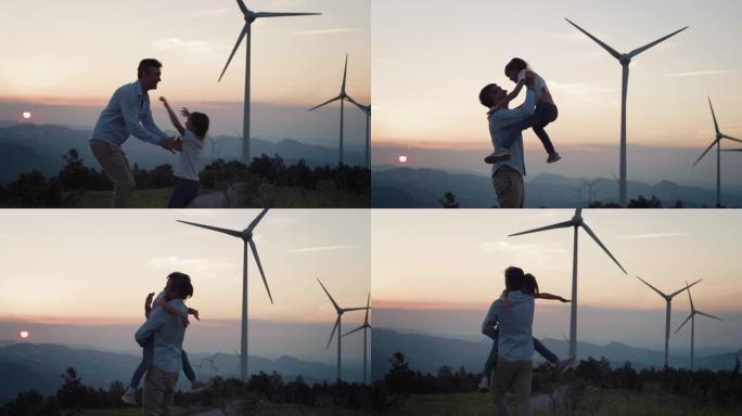 日落时，小女孩在风电场里奔跑和跳跃拥抱父亲的中等镜头。父母和孩子在被风力涡轮机包围的可持续环境中分享