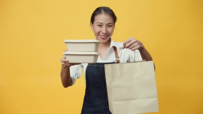 快乐微笑的亚洲女士企业主友好地看着相机里美味的食物和饮料放在纸盒里，放在黄色背景上的工艺纸袋里。