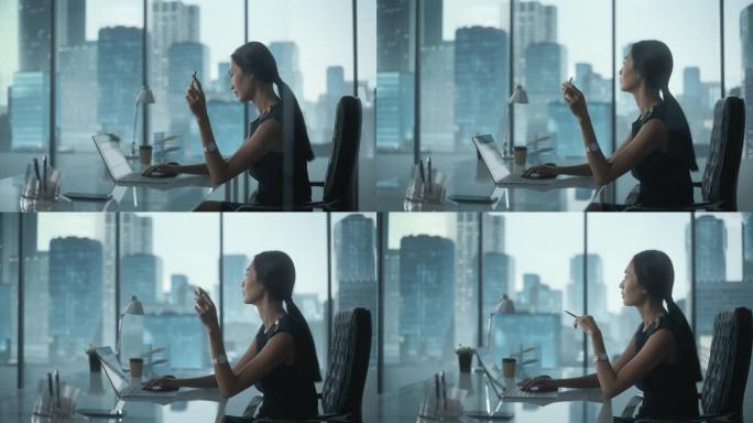 成功的女商人穿着时髦的衣服坐在现代办公室的办公桌前，使用笔记本电脑，在窗户旁边，可以看到大城市的景色