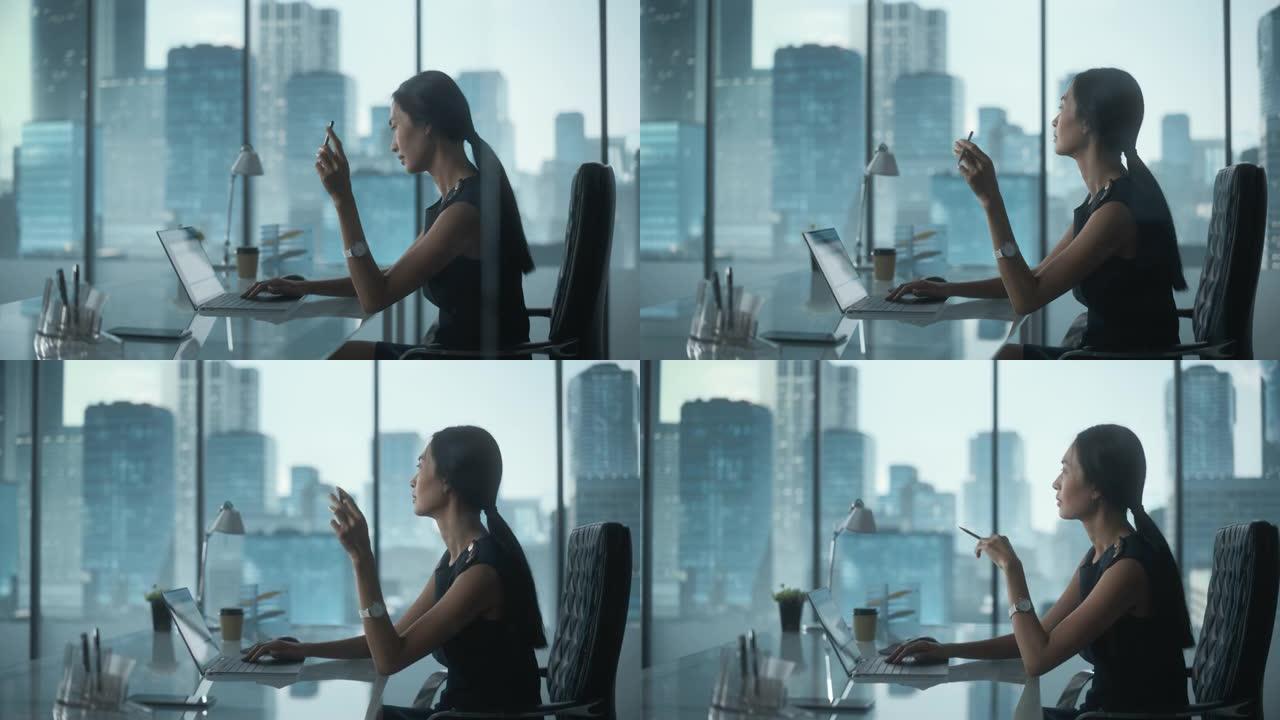 成功的女商人穿着时髦的衣服坐在现代办公室的办公桌前，使用笔记本电脑，在窗户旁边，可以看到大城市的景色