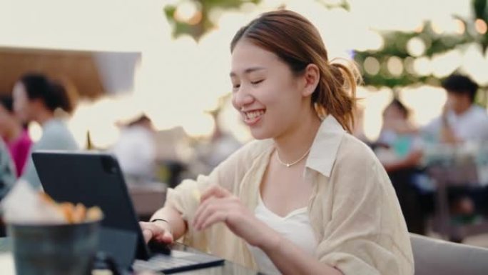 快乐的亚洲女人坐在海边的餐厅时使用数字平板电脑。
