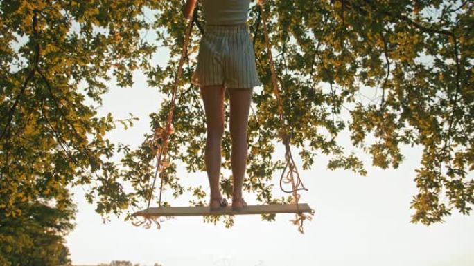 慢动作，手持镜头拍摄一个年轻的赤脚女孩，站在阳光充足的草地上悬挂在大树枝上的绳索秋千上