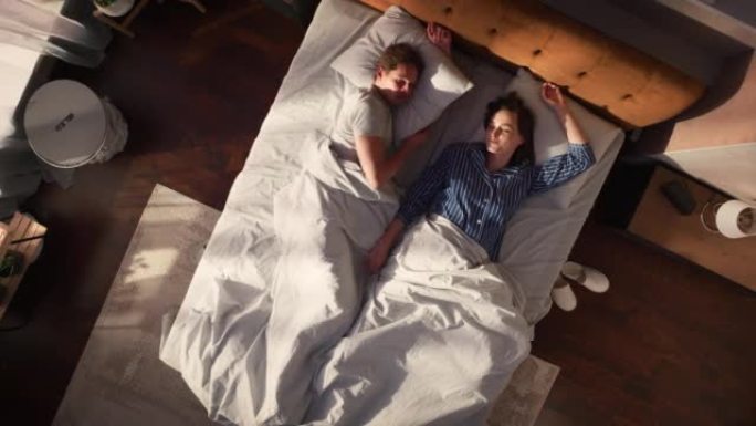 顶景公寓: 美丽的年轻夫妇睡在床上，阳光照在他们身上，他们互相看着对方，迎接新的晴天。两口之家的快乐