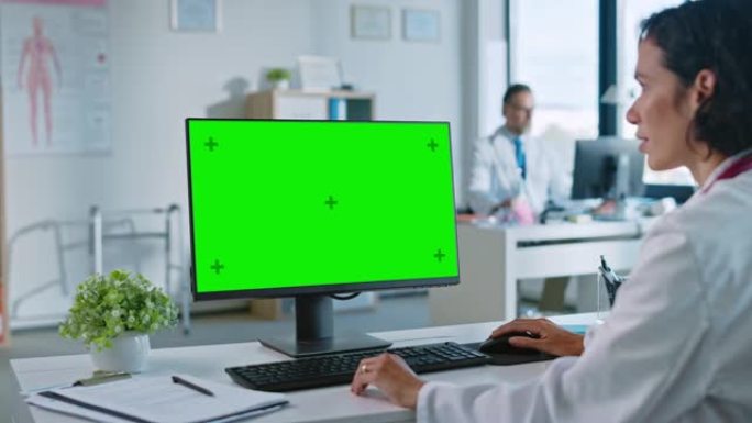 女医生正在健康诊所的绿屏模拟显示计算机上工作。穿着白大褂的助理正在医院办公室的桌子后面阅读病史。