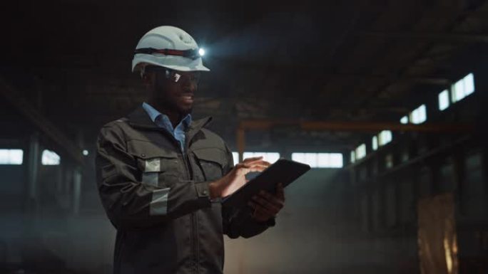 专业重工业工程师穿着安全制服和带手电筒的安全帽使用平板电脑。微笑的非洲裔美国工业专家站在金属制造仓库