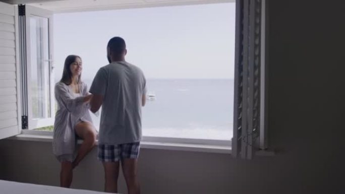年轻的浪漫夫妇在度假胜地坐在窗台旁亲吻和喝咖啡，度假时可以看到海滩景色。一个英俊的年轻人和他美丽的女