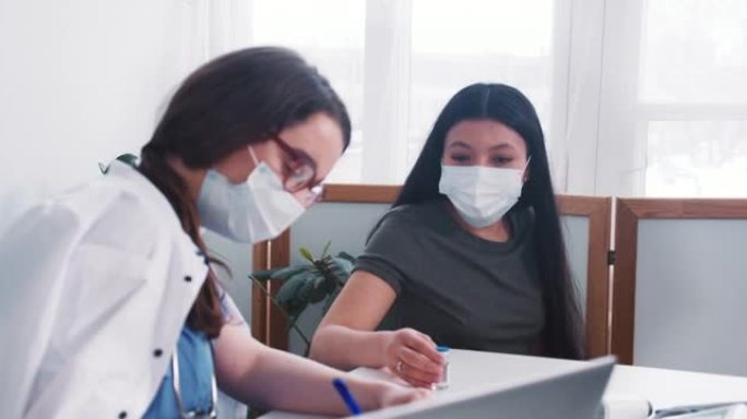 年轻的高加索医生妇女咨询戴口罩的混合种族病人妇女，在体检时拿着药瓶。