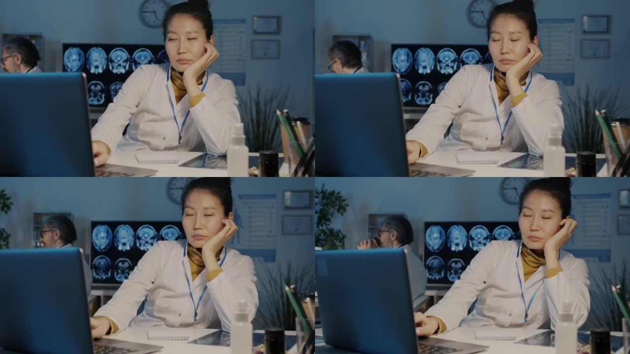 疲惫的亚洲女医生的慢动作肖像，晚上在黑暗的办公室里用笔记本电脑打盹