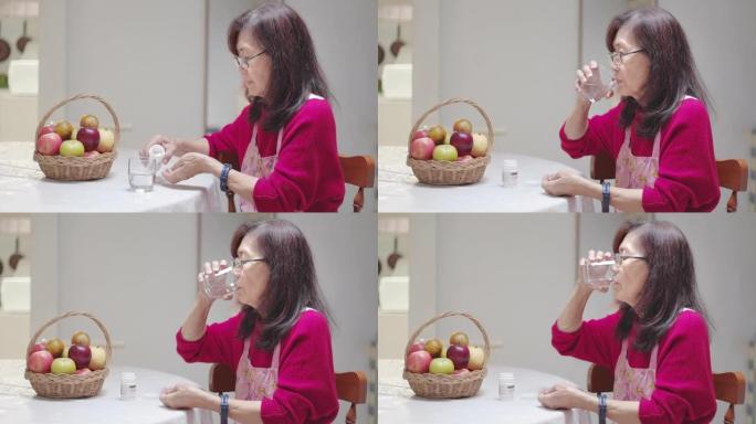 特写亚洲高级女性坐在家里的厨房里吃药或药丸，吃药后喝水