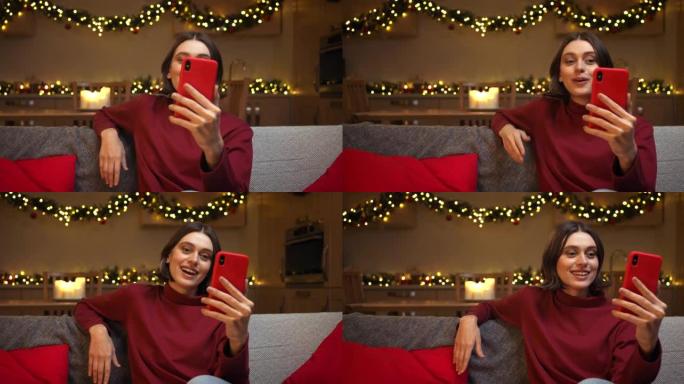 一名年轻女子通过红色智能手机上的视频通话与某人交谈，微笑着坐在舒适的圣诞节装饰厨房的沙发上