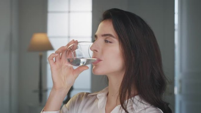 年轻的微笑女人的电影镜头正在家里的客厅用透明玻璃杯喝新鲜的纯净水。她保持身体健康，感觉健康。