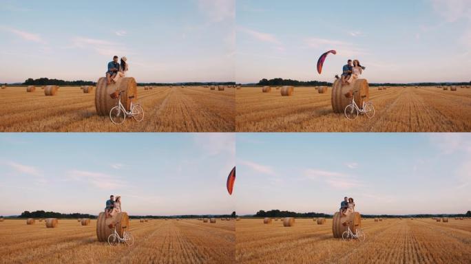 成年夫妇坐在田野上的干草捆上，滑翔伞在背景中飞行的拉出式慢动作镜头
