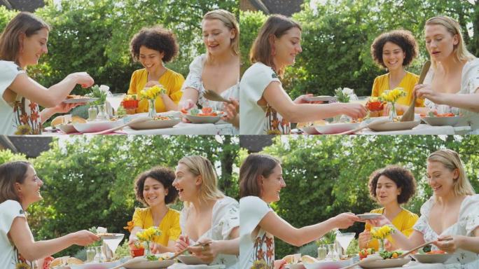 三个女性朋友坐在户外的夏季花园里，在家吃健康的饭菜，喝鸡尾酒 -- 慢动作拍摄