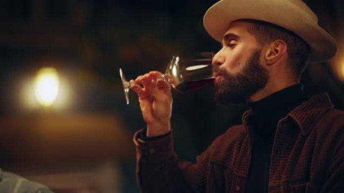 在户外露台上，一个穿着时髦衣服的英俊男子和一顶软呢帽喝着红酒的肖像。迷人的绅士享受生活，在周末的夜晚
