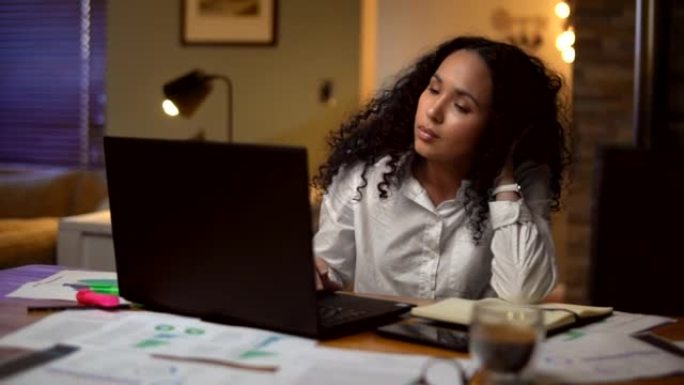 疲倦的年轻女商人坐在家庭办公室的办公桌旁看着文书工作并使用笔记本电脑。女企业家看上去精疲力尽，并试图