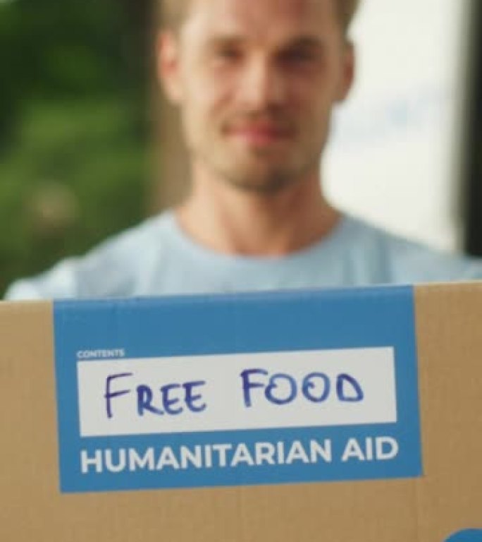 一个快乐的年轻男性志愿者的垂直肖像。穿着蓝色t恤的英俊白人男子微笑着，向观众伸出人道主义援助箱。捐赠