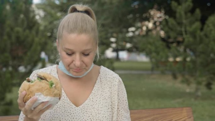 带防护口罩的SLO MO女人在公园吃汉堡并检查电话