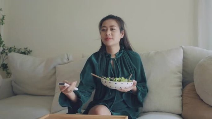 年轻快乐的亚洲女人坐在客厅的沙发上吃健康的沙拉。