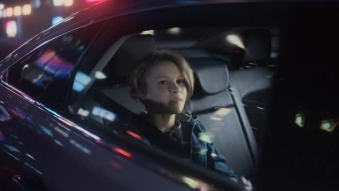 兴奋的小男孩坐在汽车后座上，晚上通勤回家。快乐而微笑的年轻孩子被带霓虹灯的城市街道上带回家。电影镜头
