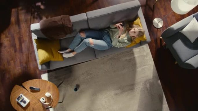 公寓: 年轻女子使用智能手机躺在客厅的沙发上。女孩在沙发上放松很开心。自由职业者在家远程工作，做电子