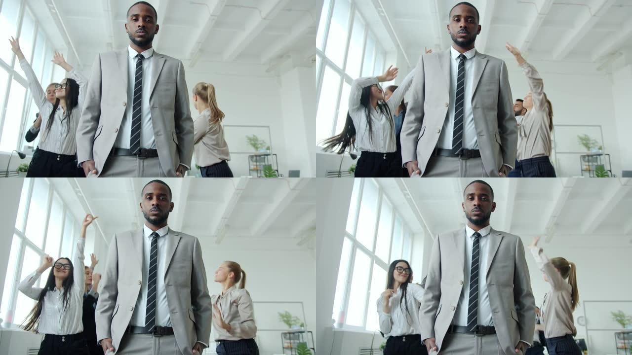 严肃的非洲裔美国人站在办公室，快乐的人在背景中跳舞的慢动作肖像