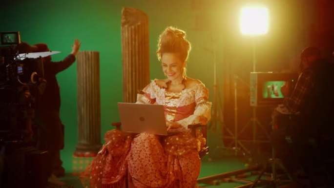 在古装戏剧电影中: 美丽的微笑女演员穿着文艺复兴时期的连衣裙，坐在椅子上，使用背景为绿屏的笔记本电脑