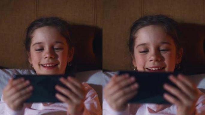 垂直屏幕: 小快乐女孩睡觉前在床上看智能手机上的娱乐视频。可爱的孩子在社交媒体网站上在线观看卡通和有