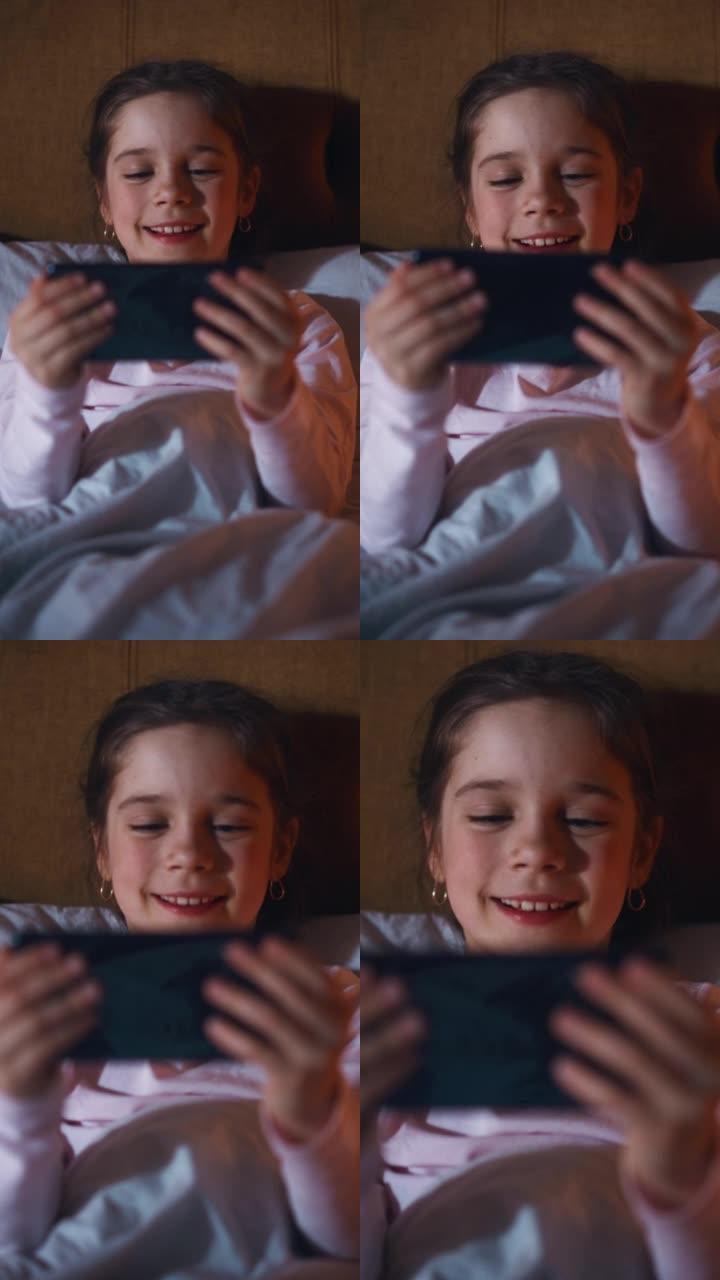垂直屏幕: 小快乐女孩睡觉前在床上看智能手机上的娱乐视频。可爱的孩子在社交媒体网站上在线观看卡通和有