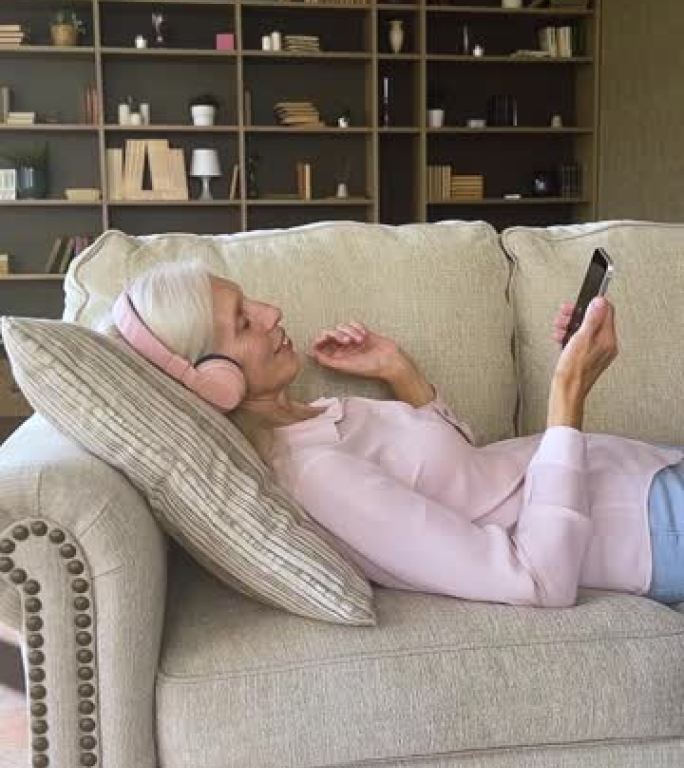 和平的高级女性戴着耳机在沙发上用手机放松