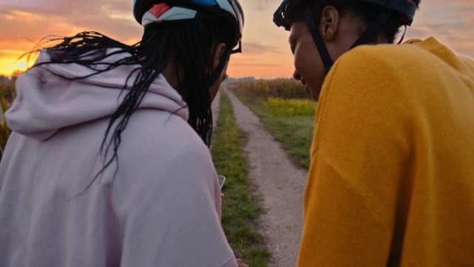 手持慢动作的两名成年女性看着智能手机，沿着草地土路骑行，朝着日落