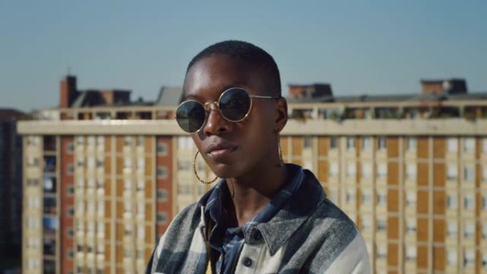 年轻时尚的黑人女性戴着太阳镜站在城区的屋顶上的电影镜头。自信的非裔美国女性看着镜头，挑战刻板印象，拥