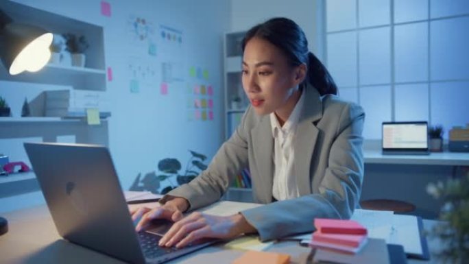 年轻的亚洲开朗的职业女商人晚上坐在桌子上，拿着笔记本电脑在现代办公室进行在线营销。