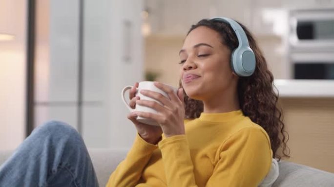 4k视频片段，一名年轻女子在家中使用耳机并在沙发上喝咖啡