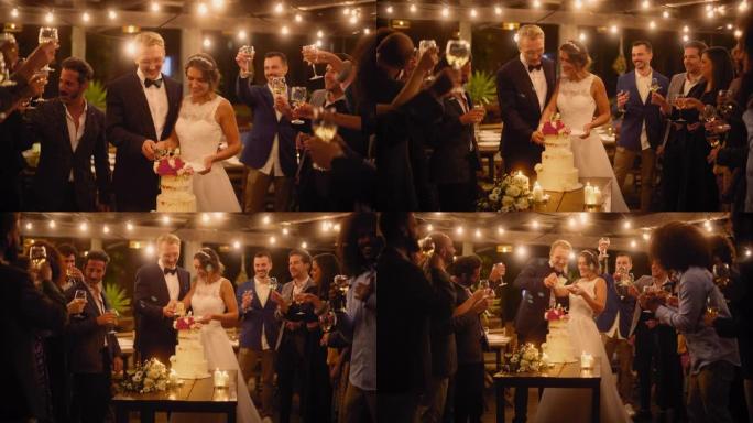 美丽的新娘和新郎在晚宴上与多民族朋友庆祝婚礼。已婚夫妇站在餐桌上，亲吻并切婚礼蛋糕。