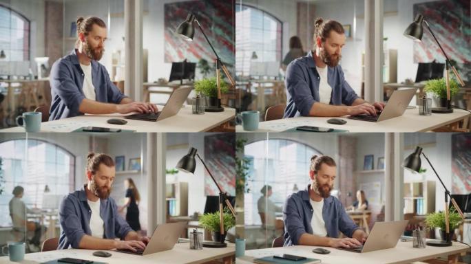 英俊的长发大胡子经理坐在创意办公室的办公桌前。年轻的时尚男人在营销机构使用笔记本电脑。在后台工作的同