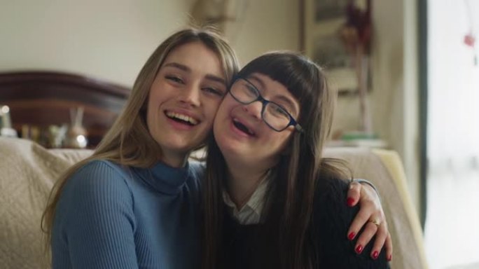 真实的快乐年轻女子拥抱她患有唐氏综合症的妹妹，在家对着镜头微笑。爱情、家庭、残疾人、医疗保健、残疾、
