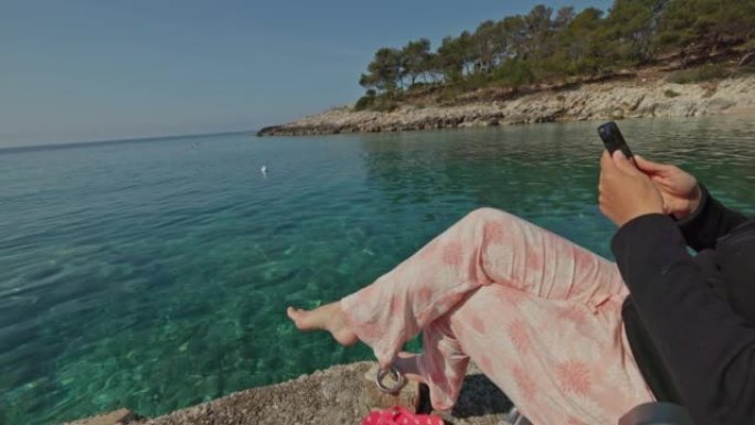 穿着睡衣的女人坐在海边岩石上的露营椅上，用手机范生活