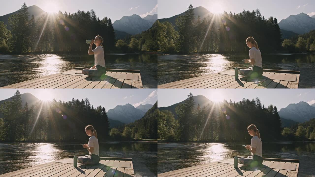 金发碧眼的女人，坐在湖前的瑜伽马特上，从训练、饮酒和使用手机中休息一下