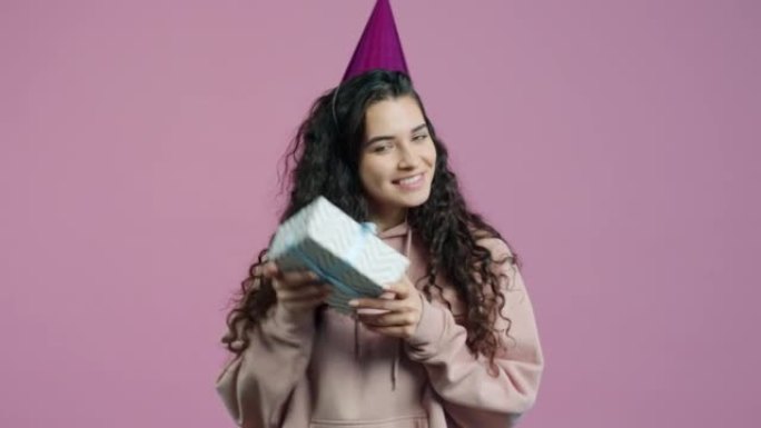 粉色背景上戴着党帽摇晃礼品盒的快乐年轻女子肖像