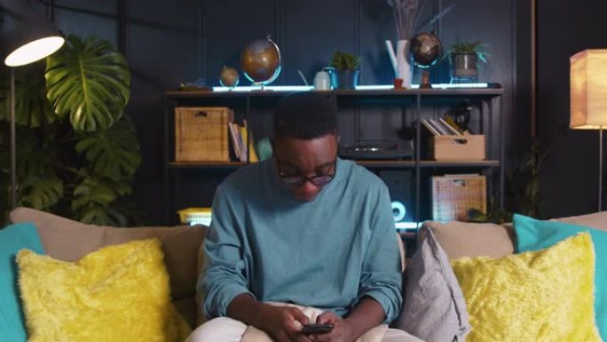 移动游戏快乐的年轻z世代黑人男学生沉迷于在家中沙发上使用智能手机在线玩游戏。