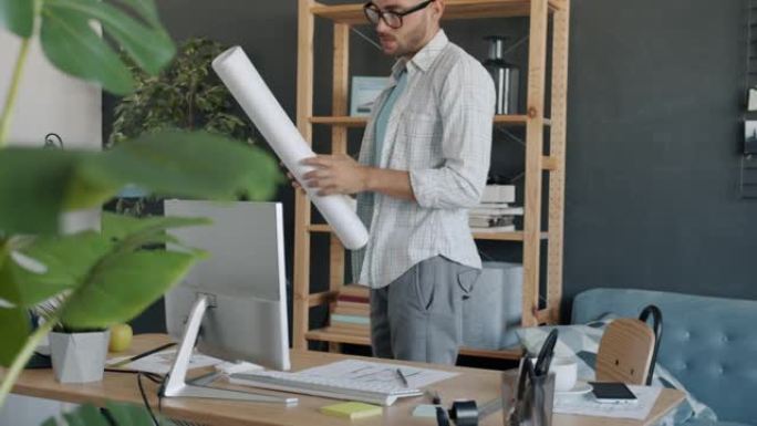 认真的男性建筑师的肖像在纸上研究蓝图并看着在公寓中工作的计算机屏幕
