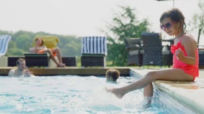 女孩坐在室外游泳池的边缘，在家庭暑假里把脚溅在水里 -- 慢动作拍摄
