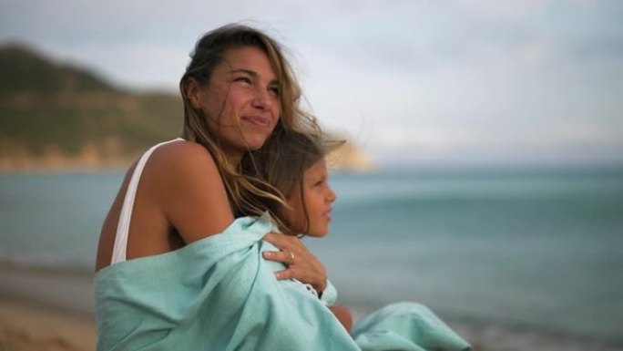 一个年轻无忧无虑的快乐微笑的母亲和被拥抱在条纹封面下的小女儿的真实照片在日落时在海边享受着他们的时光