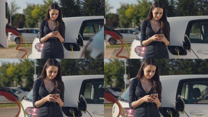 美丽的年轻女子在停车场给电动车充电时使用手机