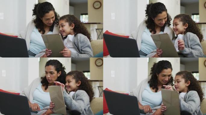 快乐的混血母女坐在沙发上，玩得开心，用笔记本电脑和平板电脑