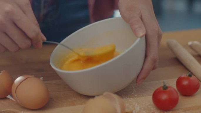 年轻的亚洲女厨师的手将鸡蛋搅拌到陶瓷碗中，在家里的厨房桌子上的木板上用蔬菜烹饪煎蛋。