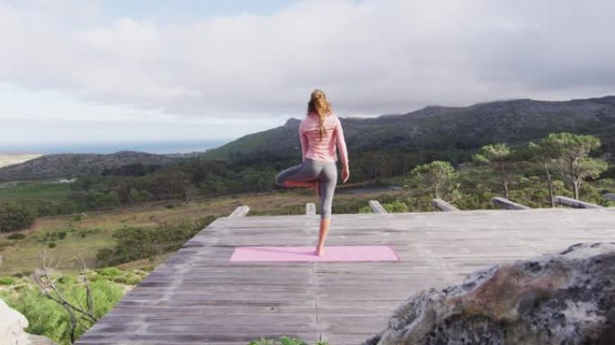 白人妇女练习瑜伽的后视站在一条腿上，在山腰的甲板上伸展