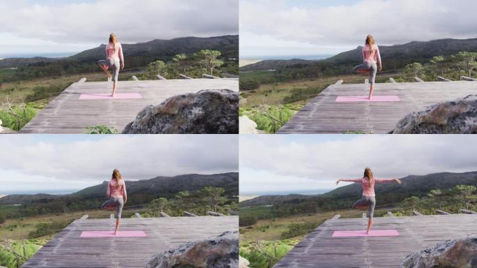 白人妇女练习瑜伽的后视站在一条腿上，在山腰的甲板上伸展