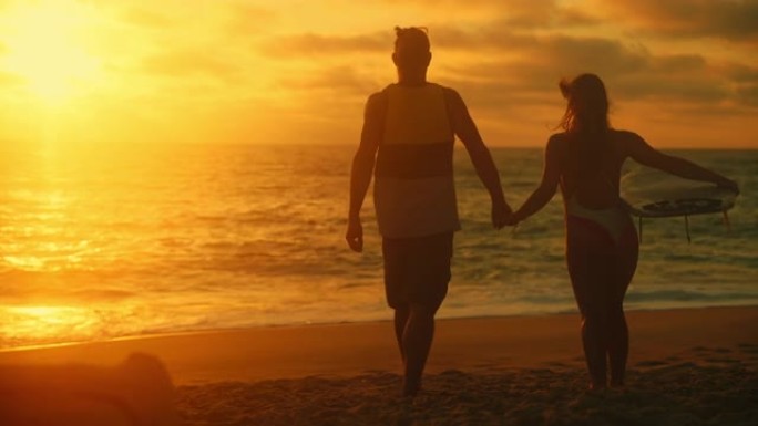 夫妻，女人和男人在日落时从海滩走到海边，手牵着手，女人在手臂下背着冲浪板，后视