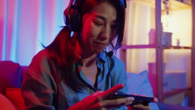 快乐亚洲女孩游戏玩家戴着耳机比赛在线视频游戏与智能手机兴奋的谈话与朋友坐在沙发上在彩色霓虹灯在家里的